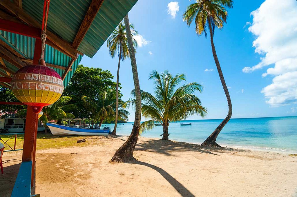 Go on...spend a little time at the beach. You know you want to. Destinations Worth Dreaming Image: A beach scene shows a boat docked in the sand; a section of a cabana; palm trees, and blue skies overlooking serene bright blue water.