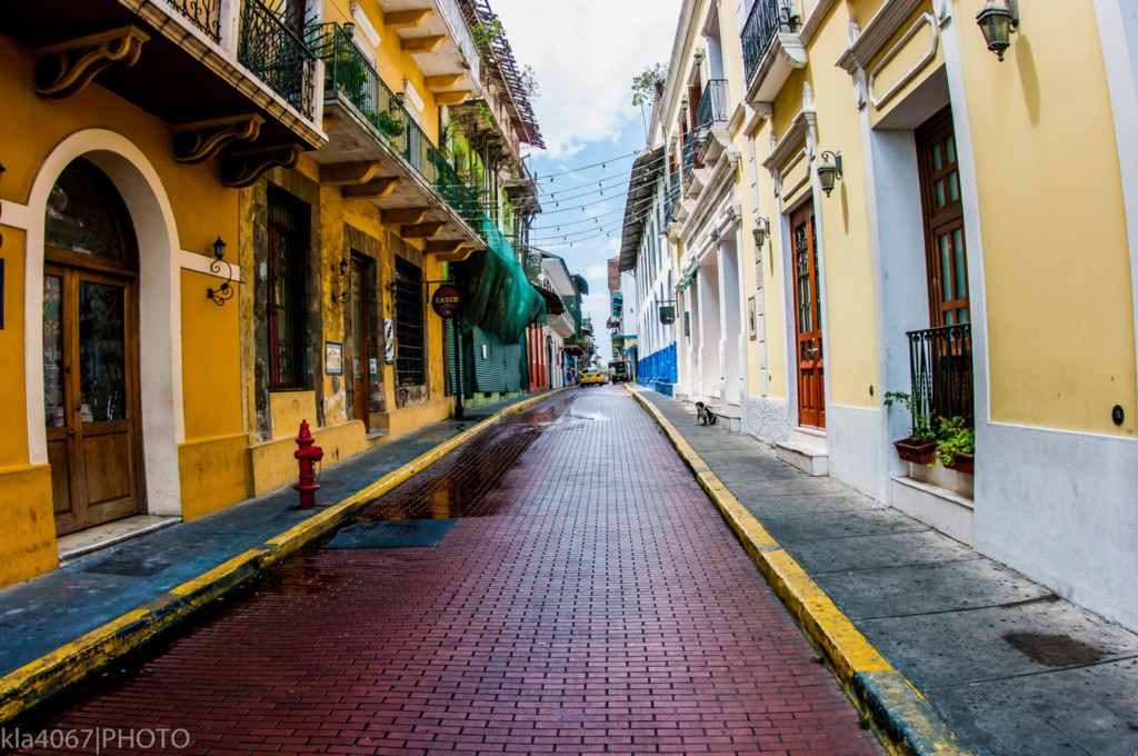 Colourful buildings always seem to have an added charm. Architecture In Panama Image: A narrow city street is lined with buildings on both the left and right. We can see shades of mustard yellow, pale lemon, turquoise, and marine blue gracing their faces. Many have wrought iron balconies.