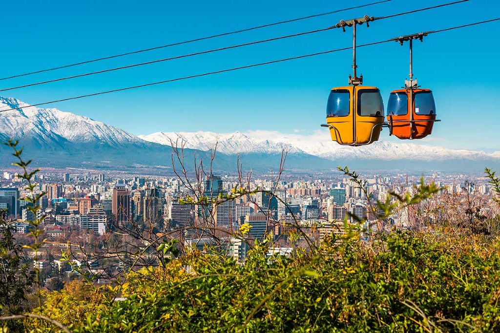 Enjoy your aerial tour, but remember, there's a lot to explore down there! Destinations Worth Dreaming Image: An enclosed aerial tram has two pods—one yellow, one orange. They are sailing above the city of Chile; a snow-capped mountain range sits in the background, the city skyline is below, and greenery graces the foreground.