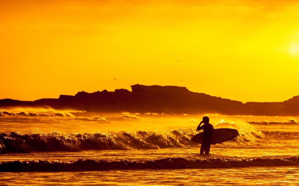 In light like this it's almost like you're surfing on a sea of gold. Destinations Worth Dreaming Image: A surfer stands in the ocean observing waves. We see little more than the silhouette because the sunlight is faded, and bathing everything in shades of orange and gold.