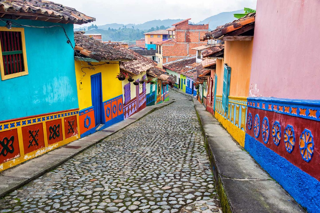 Welcome to beautiful and colourful Colombia! Destinations Worth Dreaming Image: Photograph of a camera looking down a cobblestone street; to the left and right are brightly coloured buildings with tiled roofs.