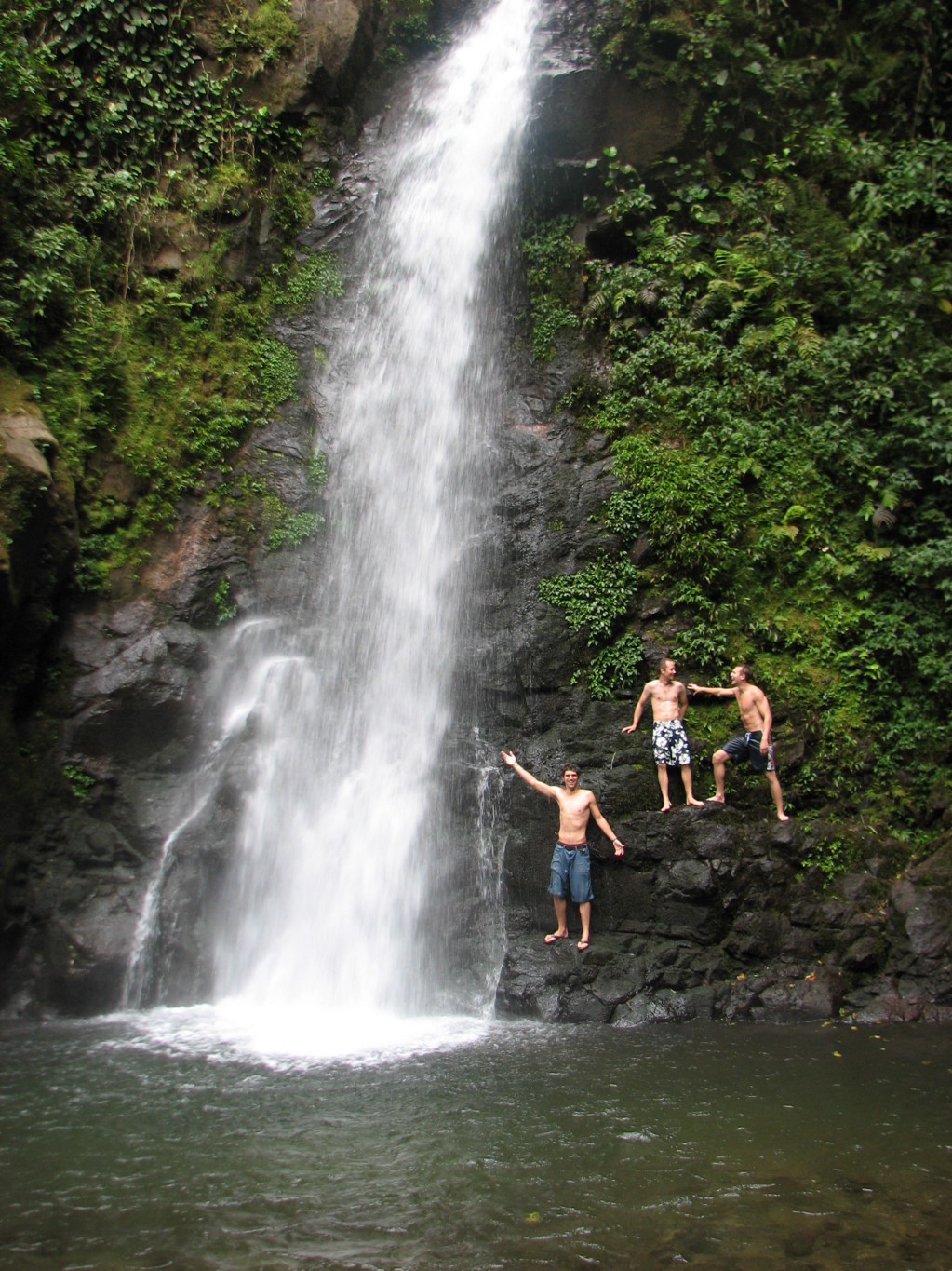 Behold this magnificent swimming hole! Swimming Holes Image: Three young men in swim shorts pose near the foot a swimming hole that has a waterfall pouring into it.