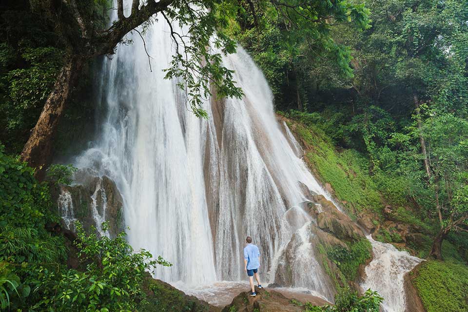 Enjoy the sound, sight, and mist of a cascading waterfall. Destinations Worth Dreaming Image: A male traveller wearing dark-blue shorts and a light-blue shirt stands on a rock observing a waterfall as it rushes and breaks down a slope in—streaming into white channels.