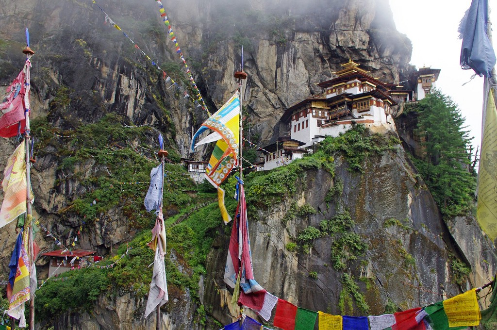Welcome to Bhutan. Destinations Worth Dreaming Image: Colourful flags and fabrics are suspended throughout as we see buildings leading up a mountainside.