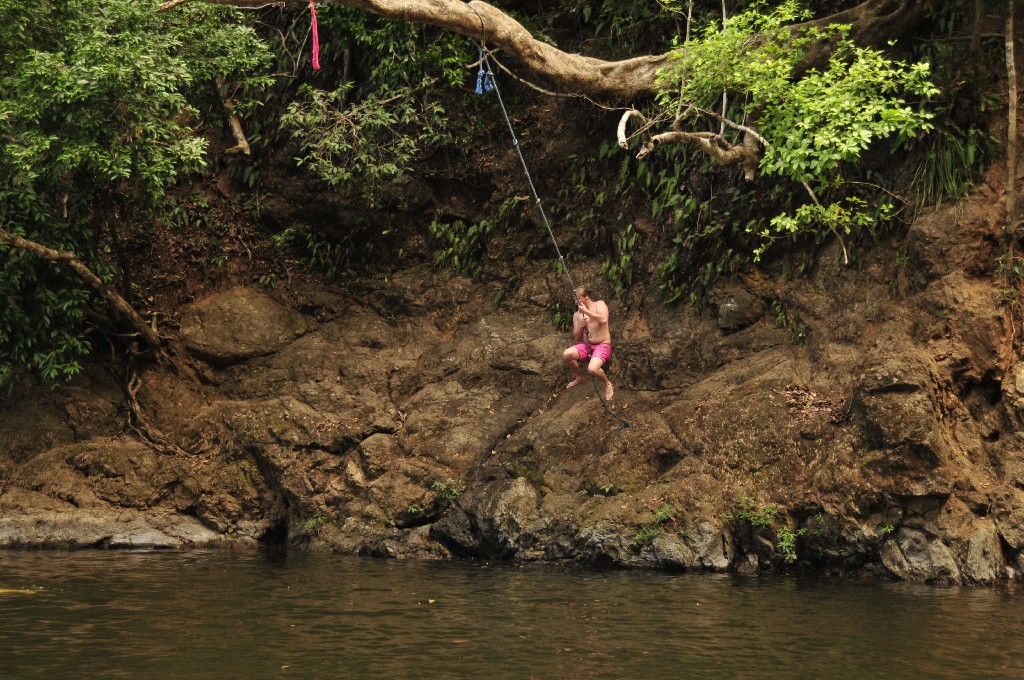 Prepare to make a splash! Swimming Holes Image: A young man in red swim shorts prepares to jettison himself off of rocks to swing on a rope suspended from a tree branch, and plunge into the water below.