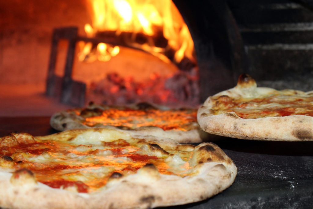 Pizza pie in paradise? Check! Costa Rican Foods Image: Three pizzas sit in the foreground whilst the open mouth of the pizza oven glows with fire in the background.