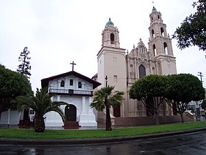Old churches, even when not official museums, are oft overlooked points of interest. Cuba Unusual Museum Image: A photograph shows a smaller white chapel with a black roof, and the larger main cathedral on a tree-lined street.