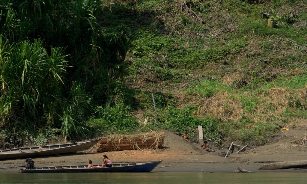 Go on holiday—see the world and its people. Rainforests & Natural Wonders: Manu National Park. Seen are people who may be members of an indigenous tribe sitting on the edge of a shore, and in a long narrow boat.