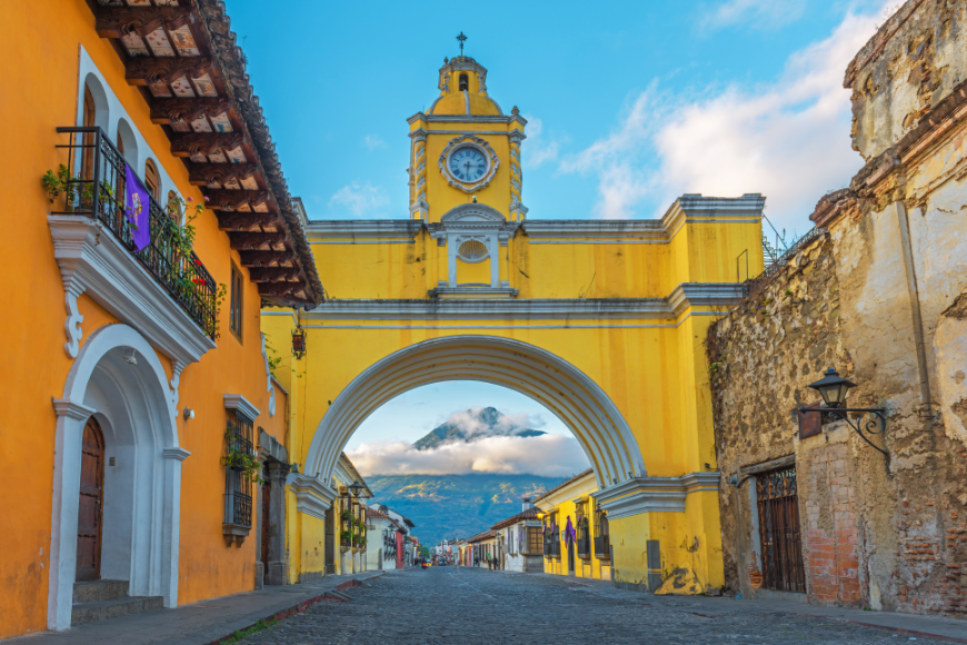 The colorful elegance of Guatemala's Spanish-Colonial architecture. Belize and Guatemala Itinerary Image: Yellow Spanish-Colonial arch on a street in Antigua, with mountains seen through the archway.