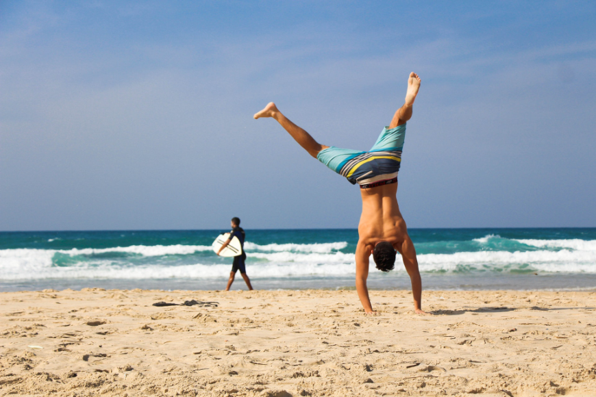 Rediscover life and joy. Moving to Belize Image: A man in striped swim shorts does a cartwheel in the sand, while a surfer in a wetsuit approaches the water in the background.