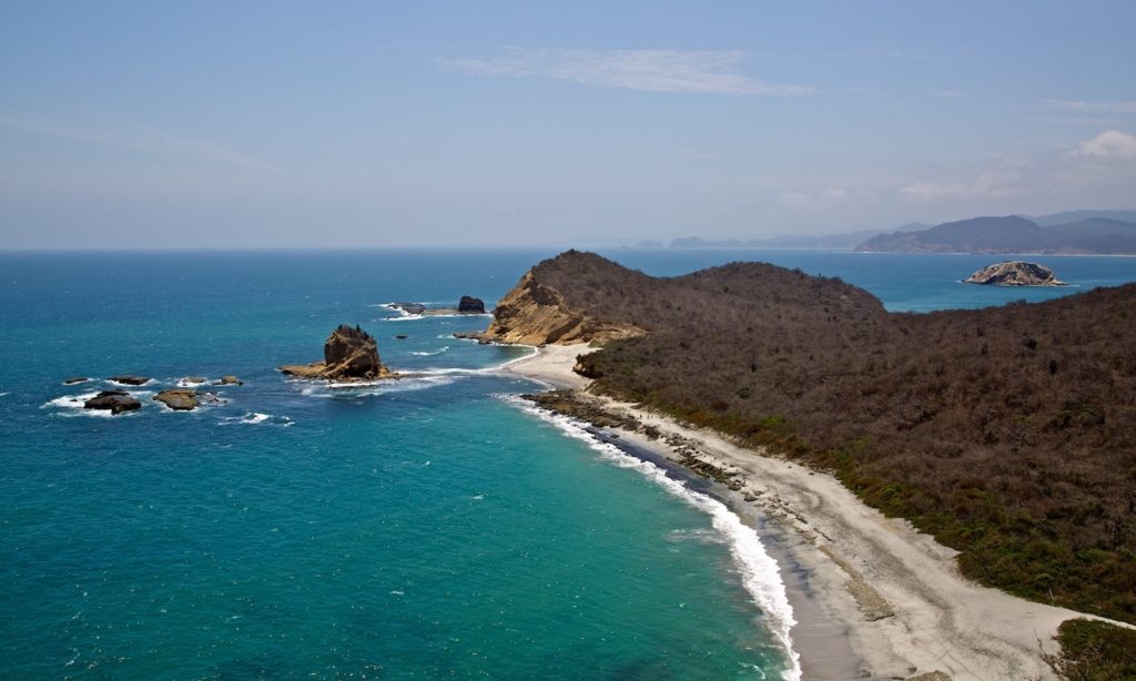 Deceptively calm in this photo, the Ecuadorian shoreline offers primo waves. Best Time To Visit South America Image: A beautiful curving coastline—green and blue waters, sandy shores, and brown and green rocky, bushy landscapes.