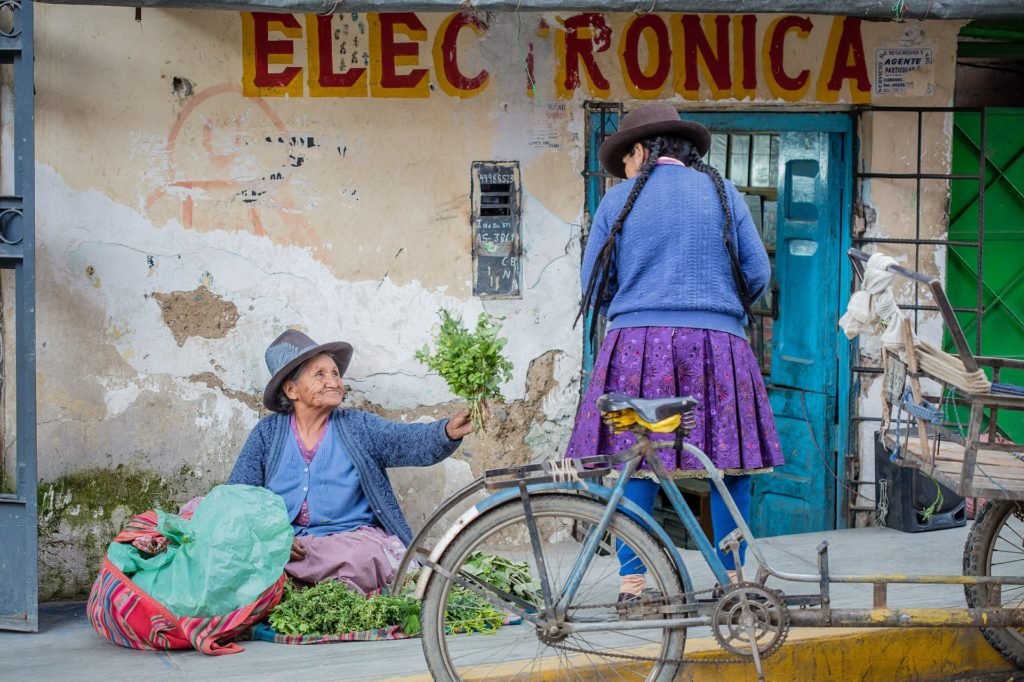 Experience the world in all its beauty and kindness. Best Time To Visit South America Image: An elder Peruvian woman sits on a sidewalk and proffers a bundle of fresh greenery to a younger woman.