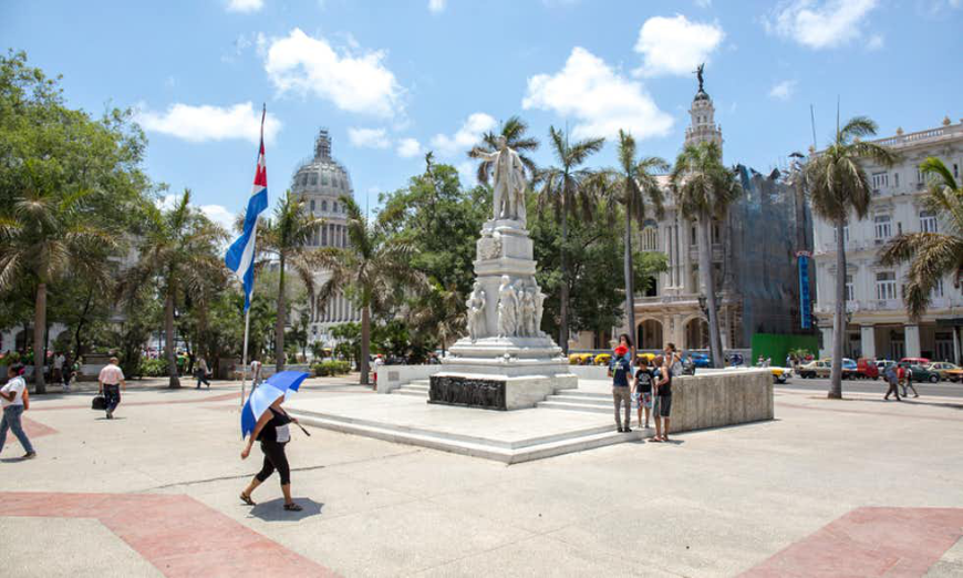 Simply walking around is a great way to get out there and explore this vibrant destination! Cuban Homestays Image: A woman walks through a plaza under an umbrella. There is a statue and a Cuban flag.