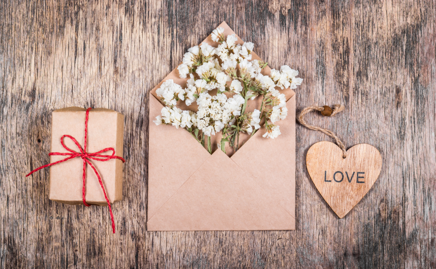 Recycled paper can be simple and elegant, and wedding favours can be crafted to last. Eco-Friendly Destination Wedding Image: Brown box with a red string bow. A wooden heart with love written on it. A brown paper envelope full of white flowers.