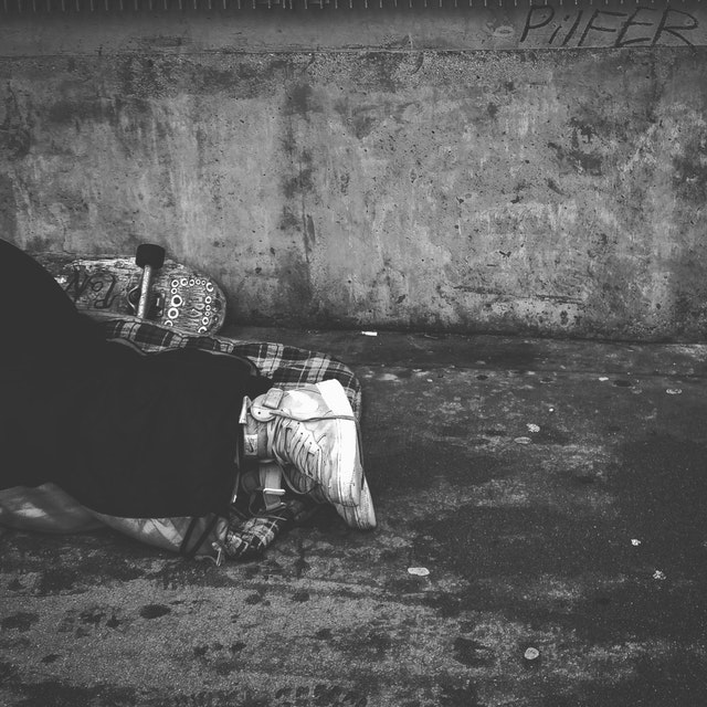 Think it's just developing countries where people don't earn enough in tourism? Think again. Environmentally Sustainable Travel: A black and white photo of someone's leg's wearing black pants and white sneakers. Behind them is a skateboard. They are sleeping on the street, laying on a plaid sleeping bag.