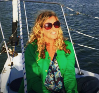 A photograph of author, Marni Switzer. Seen on a boat wearing sunglasses, and a green jacket over a printed top. Her blonde waves hang over her shoulders.