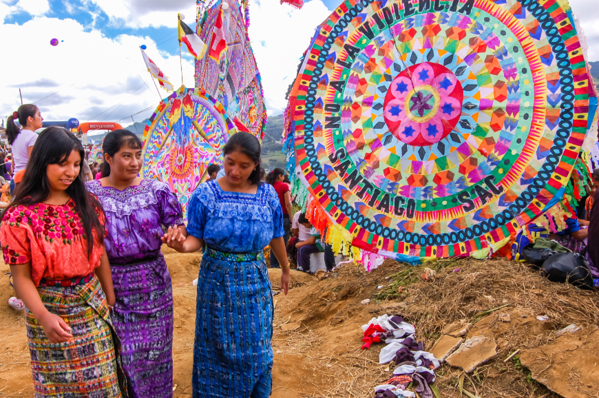 If you loved someone when they were alive, why not tout fond memories and celebrate their memory after they're gone? Mayans of Guatemala Image: Three young women in red, purple, and blue pass by colorful, oversized kites.