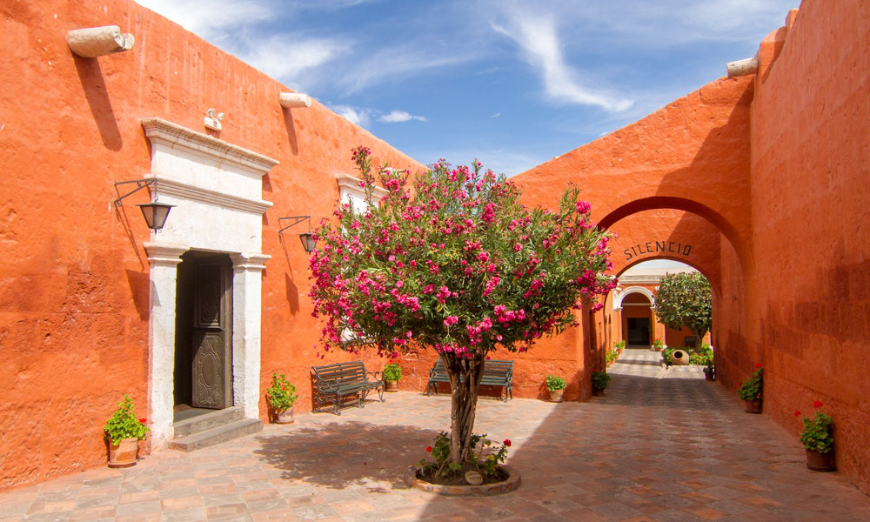 Which colour is more intense — the blue of the sky or the orange of the building? Peru Group Tour Image: An orange building and archway at the monastery; pink flowers are on a tree.