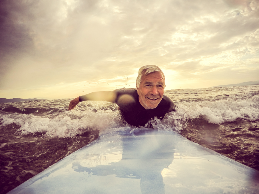 Retired? Fulfill your fantasy of surfing every day. You don't have to worry about how you'll support yourself anymore. Retiring Abroad, Moving Abroad Image: A silver haired retiree wearing a wetsuit enjoys his surf board.