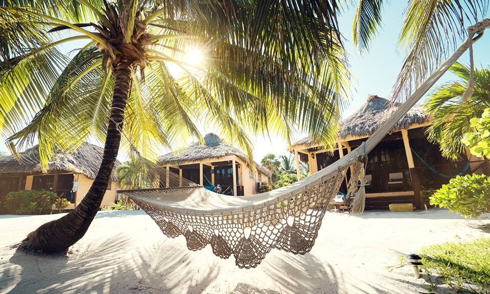 It's okay to spend your holiday napping away—so long as you have arrived at your destination first. Vacation Time Image: A crocheted cream-colored beach hammock hovers effortlessly over white sand whilst affixed to two palm trees. Three thatched roof beach huts can be seen in the background.
