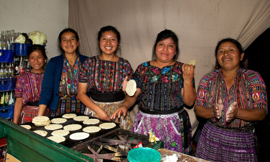 Tourism, when practiced properly, leaves both the traveler and host country better off. Sustainable Tourism Image: Women in colorful outfits prepare food at the Sololá Market in Lake Atitlán, Guatemala.
