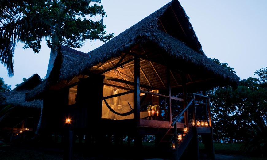 An evening so beautiful that you'll hardly miss the city lights. Sustainable Travel In Peru Image: Lanterns light the steps of a thatched roof bungalow in the midst of nature.