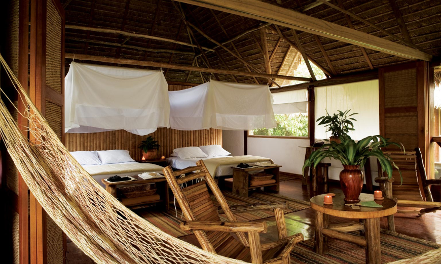 An interior view of Inkaterra Reserva Amazonica Tambopata. Sustainable Travel Practices Image: Interior view of an ecolodge room complete with canopy beds, hammock, plants, and wooden furniture.