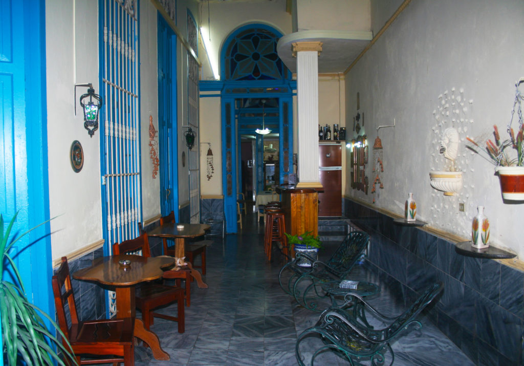 Although you can enjoy privacy at a casa particular, you can also choose to get to know your host. We recommend the latter. Legally Travel To Cuba Image: Another interior view of Casa Pardo Colonial—this time showing a room (which is almost like a very wide hallway) with bright blue door and window accents filled with tables and chairs.
