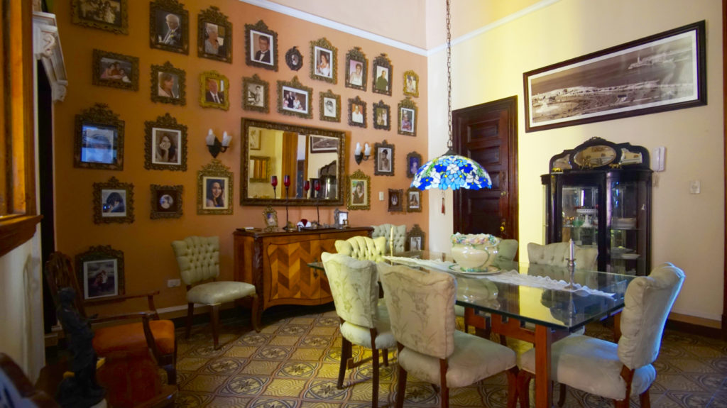 In many ways, a casa particular is similar to a bed and breakfast, or Airbnb. Legally Travel To Cuba: Another room in the casa particular Hostal del Angel; a glass dining table has six chairs, and a wall of photos, four other chairs, and additional furniture.