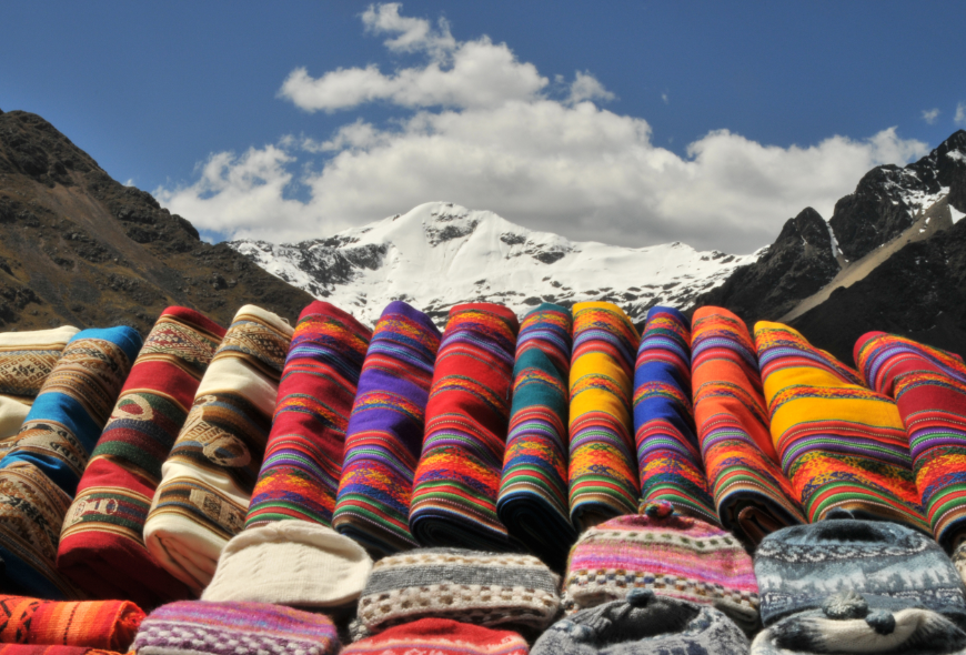 Cozy and colorful alpaca clothing is a treat for cold temperatures. Peru Souvenir Image: Colorful, traditional fabrics displayed on a stall on La Raya pass in the heart of the Inca land, Andes, Peru. Snow and glacier covered peak of Chimbuya (5600 m) visible in the background.
