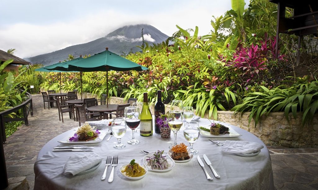 Make every evening of your anniversary or honeymoon a white tablecloth night. Unique Honeymoon Image: A delicious meal sits atop a white linen table setting, surrounded by lush and colorful foliage. A volcanic view is in the background.