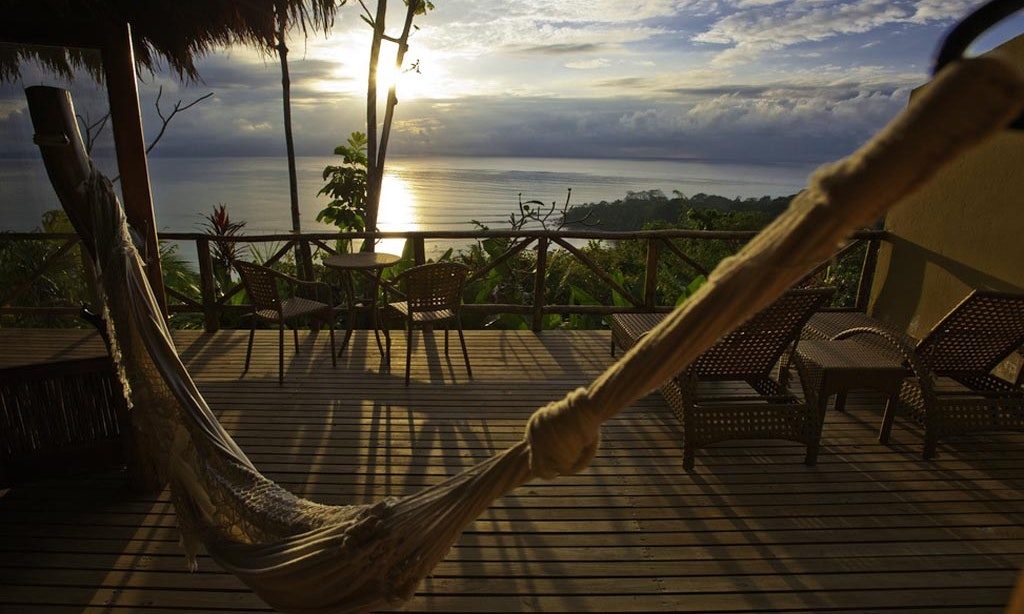 Whether you sip wine or hot chocolate, the view will remain the same—exquisite. Unique Honeymoon Image: Views of a hammock, bistro set, and lounge chairs overlook a sloping rainforest canopy and sunset.