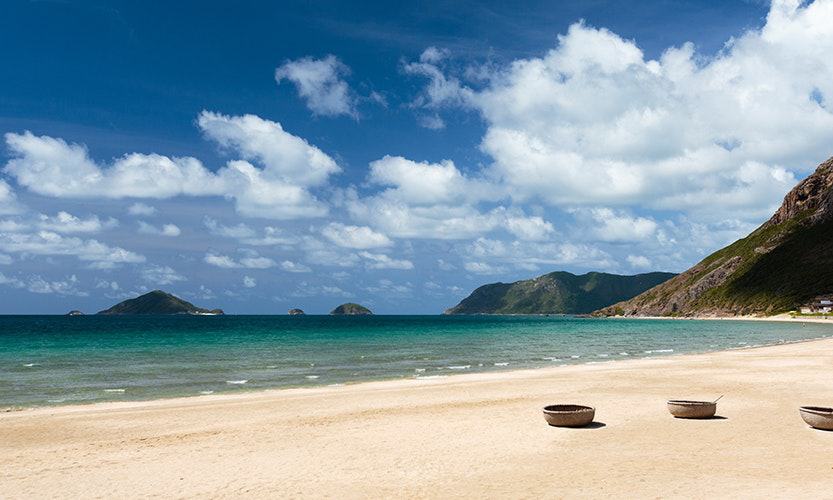 Leave your footprints in the sand, and let the tide carry them away—remember what it feels like to have fun at the beach. Vietnam Island Destinations Image: Water gently laps a beach, while blue skies and puffy white clouds grace the sky in the background.