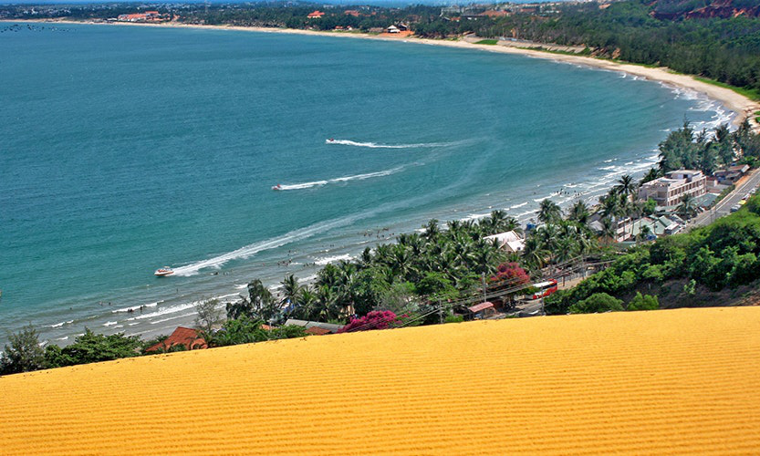 Seriously...the color of this sand is almost impossibly golden! Vietnam Island Destination Image: Golden sand dunes overlook buildings, trees, and people playing and boating in the water.