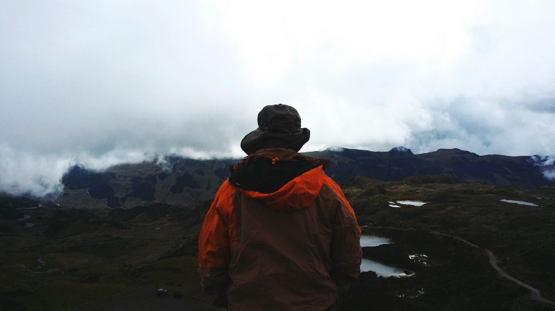 Heading to Ecuador? Pack like you're experiencing the country on foot. What To Pack Image: A person wearing a red hooded jacket and black hat (back to camera) stands gazing at a landscape of countryside, pools of water, and fog covered mountains.