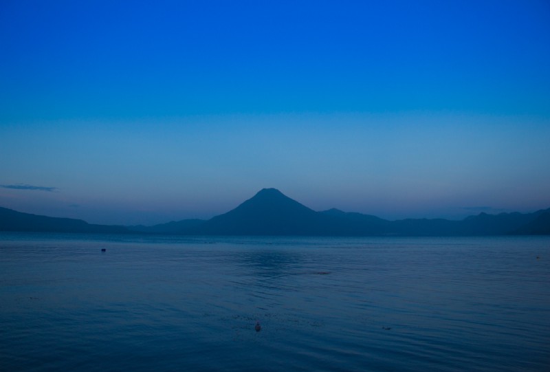 Tranquility. Serenity. Lake Atitlán. #Synonyms What To Pack Image: A lovely early-morning photograph of Guatemala's Lake Atitlán. The sky is painted in shades of dusty rose and hues of blue; gentle ripples are formed across the water.