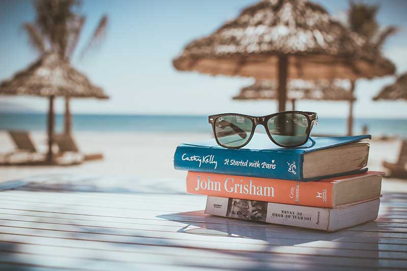 You don't realize how much you've taken beach reads for granted until you can't purchase them. What To Pack Image: A stack of three books sits on a table; a pair of black sunglasses is perched on top of them. In the background is a sandy beach, and straw beach umbrellas.