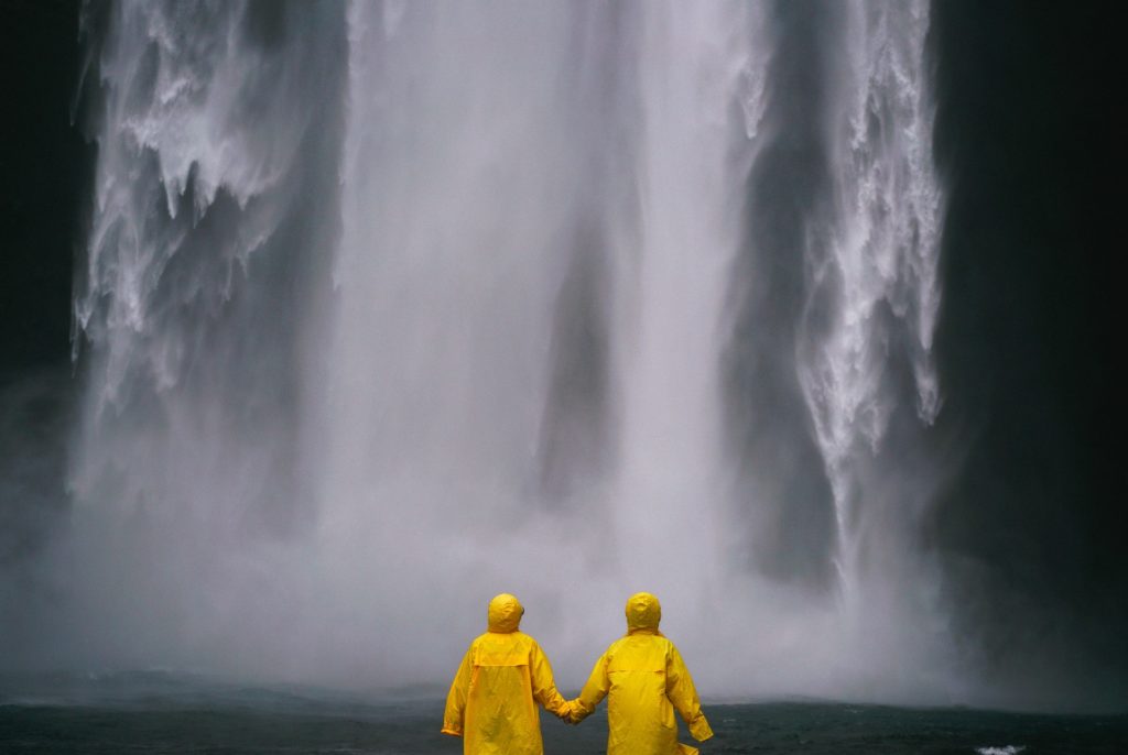 Don't let a little rain from microclimates make your holiday a washout. What To Pack Image: Two people with their backs to the camera hold hands whilst wearing yellow raincoats, and standing in front of / looking at a waterfall.