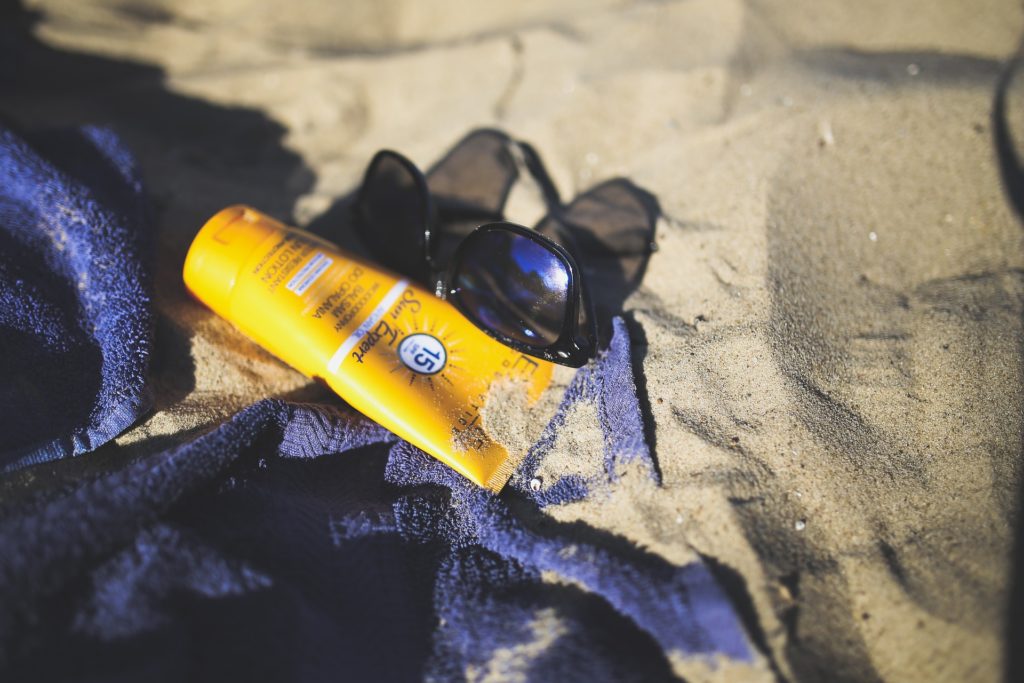 Check out those shades—chic and protective. That's the way to do it! What To Pack Image: Black sunglasses and a yellow bottle of sun protectant cream rest upon a blue towel on a sandy beach.